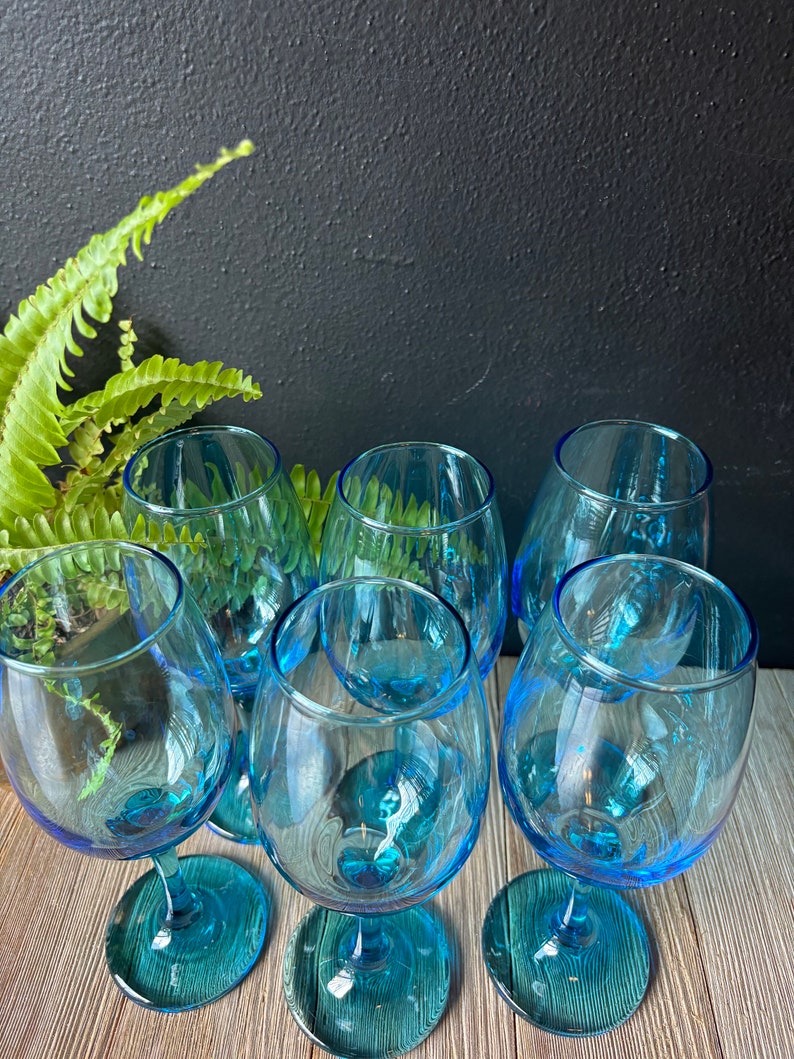 May include: Six blue glass wine glasses with a teal tint. The glasses are arranged in a circle on a wooden surface.