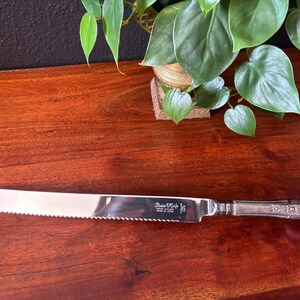 Puede incluir: Un cuchillo para mantequilla de plata con un borde dentado y un mango adornado. El cuchillo está grabado con las palabras "Butter Knife" y "Made in England".