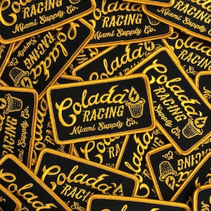 May include: A close-up of several black and gold embroidered patches with the text "Colada's Racing Miami Supply Co." in a stylized font.