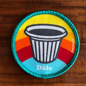 May include: A round, teal-colored patch with a rainbow design and a white coffee cup with the word "Dále" in white text.