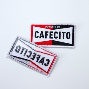 May include: Two white fabric patches with black and red trim. The top patch says "POWERED BY CAFECITO" in black text. The bottom patch says "CAFECITO" in black text, but the text is reversed.