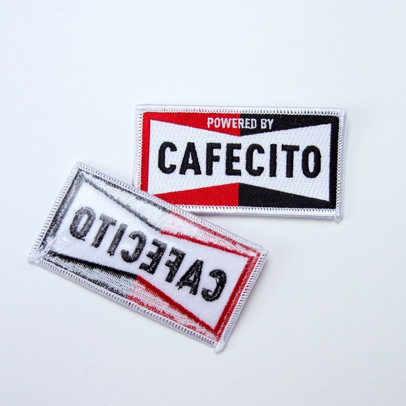 May include: Two white fabric patches with black and red trim. The top patch says "POWERED BY CAFECITO" in black text. The bottom patch says "CAFECITO" in black text, but the text is reversed.
