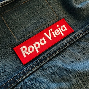 May include: A red rectangular patch with white embroidered text that reads "Ropa Vieja" sewn onto a blue denim jacket.
