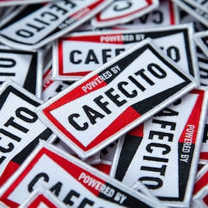 May include: A pile of white and black embroidered patches with red accents. The patches feature the text "Powered by Cafecito" in black and white lettering.