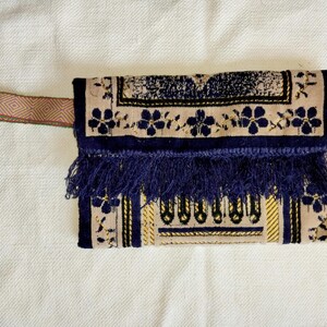 Puede incluir: Un bolso de mano beige y azul marino con un estampado floral decorativo y flecos. El bolso tiene un asa tejida con un diseño geométrico. El diseño incluye detalles dorados.