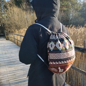 Op de afbeelding: Een rugzak met trekkoord en een kleurrijk geometrisch patroon in tinten rood, groen en bruin. De rugzak heeft witte bandjes en wordt over een zwarte jas gedragen. De achtergrond toont een houten brug en bomen.