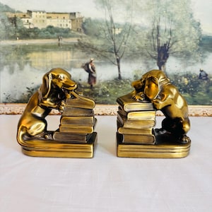 May include: A pair of brass dachshund bookends. Each bookend features a dog resting on a stack of books. The bookends have a gold finish and are set against a landscape painting.