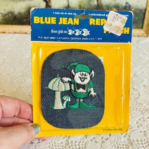 May include: Vintage blue jean patch featuring an embroidered leprechaun with a mushroom. The patch is on a yellow and blue cardboard backing with the text "BLUE JEAN REPAIR" and "THREE FISH INC."