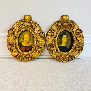May include: Two antique oval portraits in ornate, gold-colored frames. The portraits depict individuals with classical attire and features. The frames have a detailed, scrollwork design and a decorative top. The background is a white wall and a white lace tablecloth.