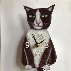 May include: A black and white cat clock with a pendulum tail. The clock face has black Roman numerals and black hands. The clock is made of wood and has a glossy finish.