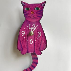 以下が含まれることがあります： ピンクの猫の形をした時計で、白い文字盤に黒い数字が書かれています。猫の尻尾は振り子です。時計の文字盤には「12、9、3、6」と表示されています。