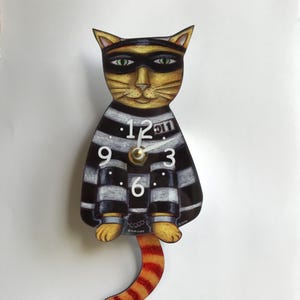 May include: A black and white striped cat clock with a black mask and orange tail. The clock face has black numbers and a white background. The clock is hanging from a hook.