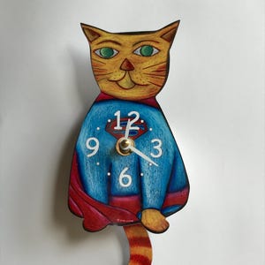 May include: A yellow cat wearing a blue Superman costume with a red cape and a clock face on its chest. The clock face has white numbers and hands. The cat's tail is orange and black.