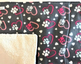 Puede incluir: Una cama reversible para mascotas con una parte superior de forro polar gris con un patrón de corazones y huellas de patas rosas y blancas. La parte inferior está hecha de un tejido sherpa suave de color crema.