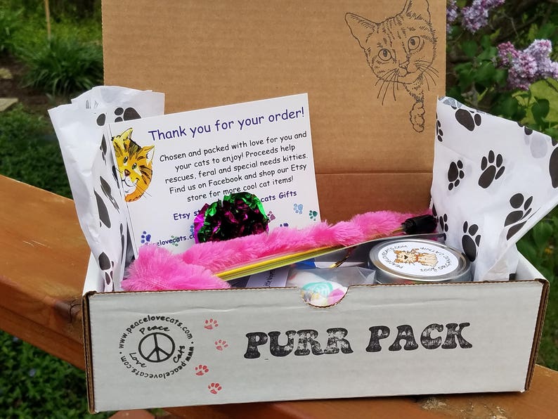 Puede incluir: Una caja de cart&oacute;n con las palabras "PURR PACK" impresas en la parte delantera. La caja est&aacute; abierta y contiene una variedad de juguetes para gatos, incluyendo una varita de plumas rosa, una lata de plata con una ilustraci&oacute;n de un gato y una bolsa blanca con huellas de patas negras. La caja tambi&eacute;n incluye una nota de agradecimiento con el texto "&iexcl;Gracias por tu pedido! &iexcl;Elegido y empaquetado con amor para ti y tus gatos para disfrutar! Las ganancias ayudan a los rescates, los gatos ferales y los gatos con necesidades especiales. &iexcl;Encu&eacute;ntranos en Facebook y compra en nuestra tienda Etsy para m&aacute;s art&iacute;culos geniales para gatos! Etsy regalos para gatos peacelovecats.com".