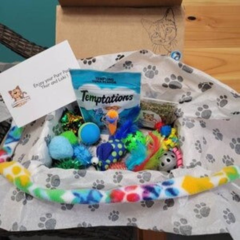 Puede incluir: Una caja de regalo llena de juguetes para gatos, incluyendo una pelota azul, una varita de plumas y un juguete de hierba gatera. La caja tambi&eacute;n incluye una bolsa de golosinas para gatos Temptations con sabor a at&uacute;n. La caja est&aacute; forrada con papel de seda con estampado de huellas de patas y una manta de franela azul y verde.