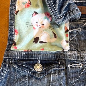May include: A denim vest with a light blue interior featuring a smiling cartoon cat with a flower crown. The vest has a button closure and a small embroidered flower detail on the pocket. The vest is made of blue denim.