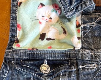 Puede incluir: Un chaleco vaquero con interior azul claro con un gato de dibujos animados sonriente con una corona de flores. El chaleco tiene cierre de botón y un pequeño detalle de flor bordada en el bolsillo. El chaleco está hecho de mezclilla azul.
