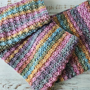 May include: Close-up of several handmade crochet items in a variety of colors, including pink, blue, green, and yellow. The items appear to be washcloths or small blankets, with a textured, striped pattern. The items are arranged on a white wooden surface.