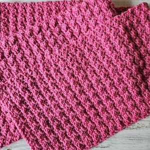 Puede incluir: Una manta de crochet rosa con un patrón de puntadas texturizadas. La manta está doblada por la mitad y colocada sobre una superficie de madera blanca.