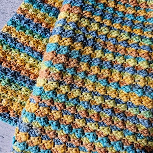 May include: Two handmade crochet squares in a textured pattern. The squares feature a mix of colors, including teal, blue, yellow, and brown. The yarn appears to be soft and the stitches are tightly woven, creating a cozy, textured look.