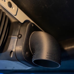 May include: A close-up of a black plastic automotive part, possibly a vent or duct, with a curved outlet. The part is mounted under a dark surface, with a light-colored bracket visible. The image is well-lit, highlighting the texture and shape.