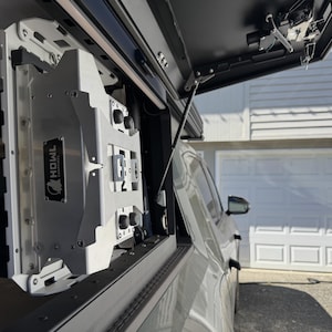 May include: A silver metal truck bed storage system with the text "HOWL COMPANY" on a black label. The system has multiple compartments, knobs, and a hinged lid. The truck bed is open, revealing the interior.