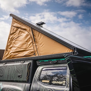 May include: A black pickup truck with a tan pop-up tent on the roof. The truck has a green and black topographic pattern wrap. The truck has a sticker that says "Jake Delger" on the back window.