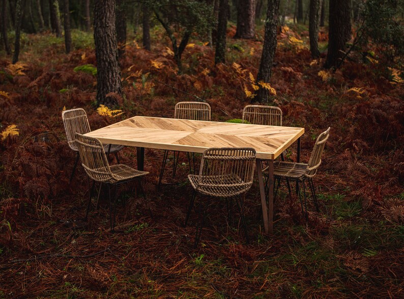 May include: A rectangular wooden dining table with a geometric pattern, set with six woven chairs. The table has dark metal legs. The scene is set outdoors in a forest with brown foliage and tall trees.