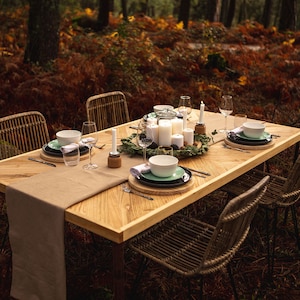 May include: A wooden dining table set for a meal in a forest setting. The table is set with white candles, plates, bowls, glasses, and silverware. Wicker chairs surround the table, and a tan runner is on the table.