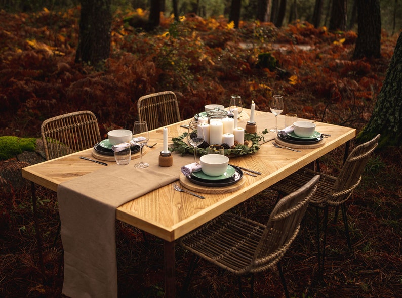 May include: A wooden dining table set for a meal in a forest setting. The table is set with white candles, plates, bowls, glasses, and silverware. Wicker chairs surround the table, and a tan runner is on the table.