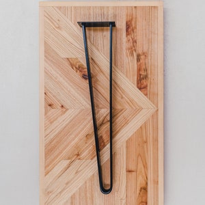 May include: A wooden table top with a geometric pattern and a black metal hairpin leg. The table top is made of light-colored wood and has a natural finish. The word "PLETINA" is written in teal at the top.