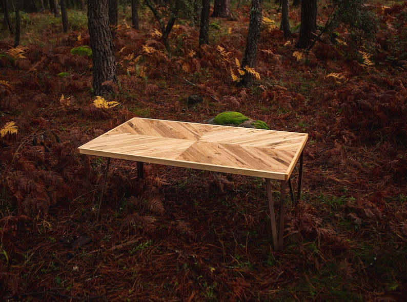 May include: A rectangular wooden table with a chevron pattern on the tabletop. The table has a light brown finish and dark metal hairpin legs. The table is approximately 60 inches long and 30 inches wide.