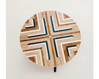 May include: Round wooden table top with a geometric pattern of brown, white, and teal wood pieces. The table has a black metal leg.