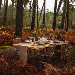 May include: A wooden dining table set for a meal in a forest setting. The table is surrounded by four woven chairs and set with plates, bowls, glasses, and candles. The forest floor is covered in brown and orange foliage.