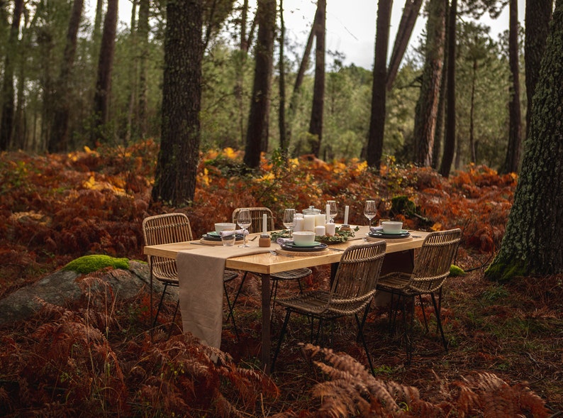May include: A wooden dining table set for a meal in a forest setting. The table is surrounded by four woven chairs and set with plates, bowls, glasses, and candles. The forest floor is covered in brown and orange foliage.