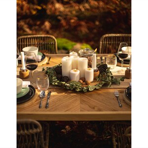 May include: A beautifully set wooden table with a centerpiece of white candles, greenery, pine cones, and walnuts. Wine glasses, plates, and silverware are arranged for a dinner setting. The scene is bathed in warm, natural light, suggesting an outdoor gathering.