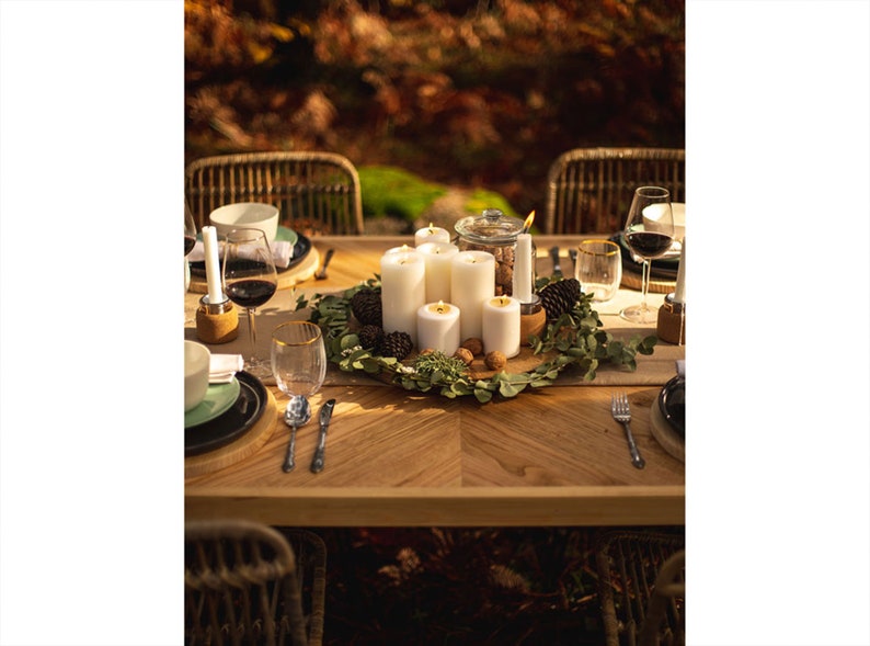 May include: A beautifully set wooden table with a centerpiece of white candles, greenery, pine cones, and walnuts. Wine glasses, plates, and silverware are arranged for a dinner setting. The scene is bathed in warm, natural light, suggesting an outdoor gathering.