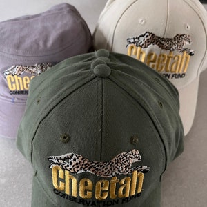 Op de afbeelding: Drie baseballpetten met een cheetah-ontwerp en de tekst "Cheetah Conservation Fund". De petten zijn in verschillende kleuren: grijs, beige en groen.