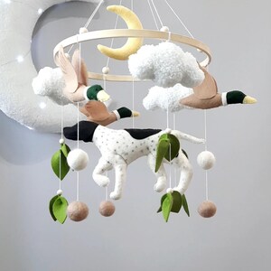 May include: A white and black spotted dog, two brown ducks, a yellow crescent moon, and white fluffy clouds hang from a wooden ring. The mobile is decorated with green felt leaves and brown felt balls.