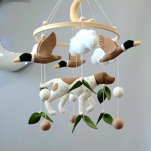 May include: A hanging nursery mobile featuring a wooden hoop frame with a central yellow crescent moon. Suspended elements include three felt mallard ducks, white pom-pom clouds, a spotted dog, green felt leaves, and small beige felt balls.