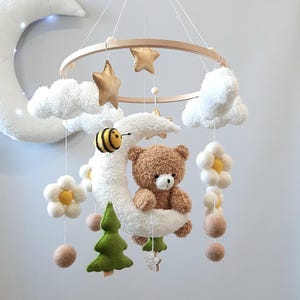 May include: A white and brown teddy bear sits on a white crescent moon with a yellow and black bee. The moon is hanging from a wooden ring with white clouds, green trees, yellow flowers, and gold stars.
