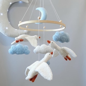May include: A white wooden baby mobile with a crescent moon, clouds, and geese. The geese are made of white fluffy fabric with orange beaks and feet. The clouds are blue and white.