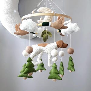 May include: A hanging nursery mobile featuring a central felt dog with brown and white spots, surrounded by hanging felt elements including green pine trees, brown ducks with green heads, white clouds, and small felt balls suspended from a wooden ring.