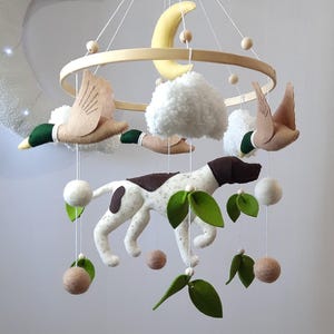 May include: A wooden baby mobile with a brown and white dog, two brown and green ducks, a white fluffy cloud, a yellow crescent moon, and green felt leaves. The mobile is hanging from a wooden ring.