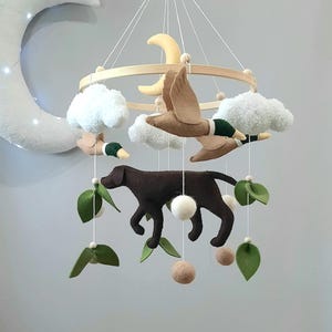 May include: A handcrafted baby mobile featuring a wooden ring with felt elements. The mobile includes brown ducks, white clouds, yellow moons, a brown dog, green leaves, and beige pom-poms. The mobile hangs from white strings.