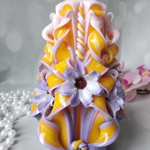 May include: A hand-carved candle with a yellow and purple striped design. The candle is decorated with white and purple flowers and has a twisted pink and white center.