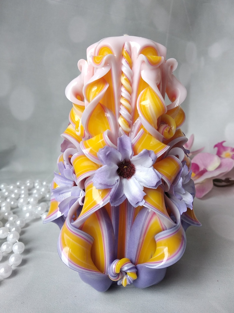 May include: A hand-carved candle with a yellow and purple striped design. The candle is decorated with white and purple flowers and has a twisted pink and white center.