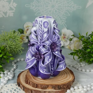Puede incluir: Una vela tallada a mano de color morado y blanco con un dise&ntilde;o floral. La vela est&aacute; sentada sobre una base de madera.
