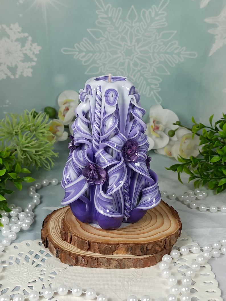 Puede incluir: Una vela tallada a mano de color morado y blanco con un dise&ntilde;o floral. La vela est&aacute; sentada sobre una base de madera.
